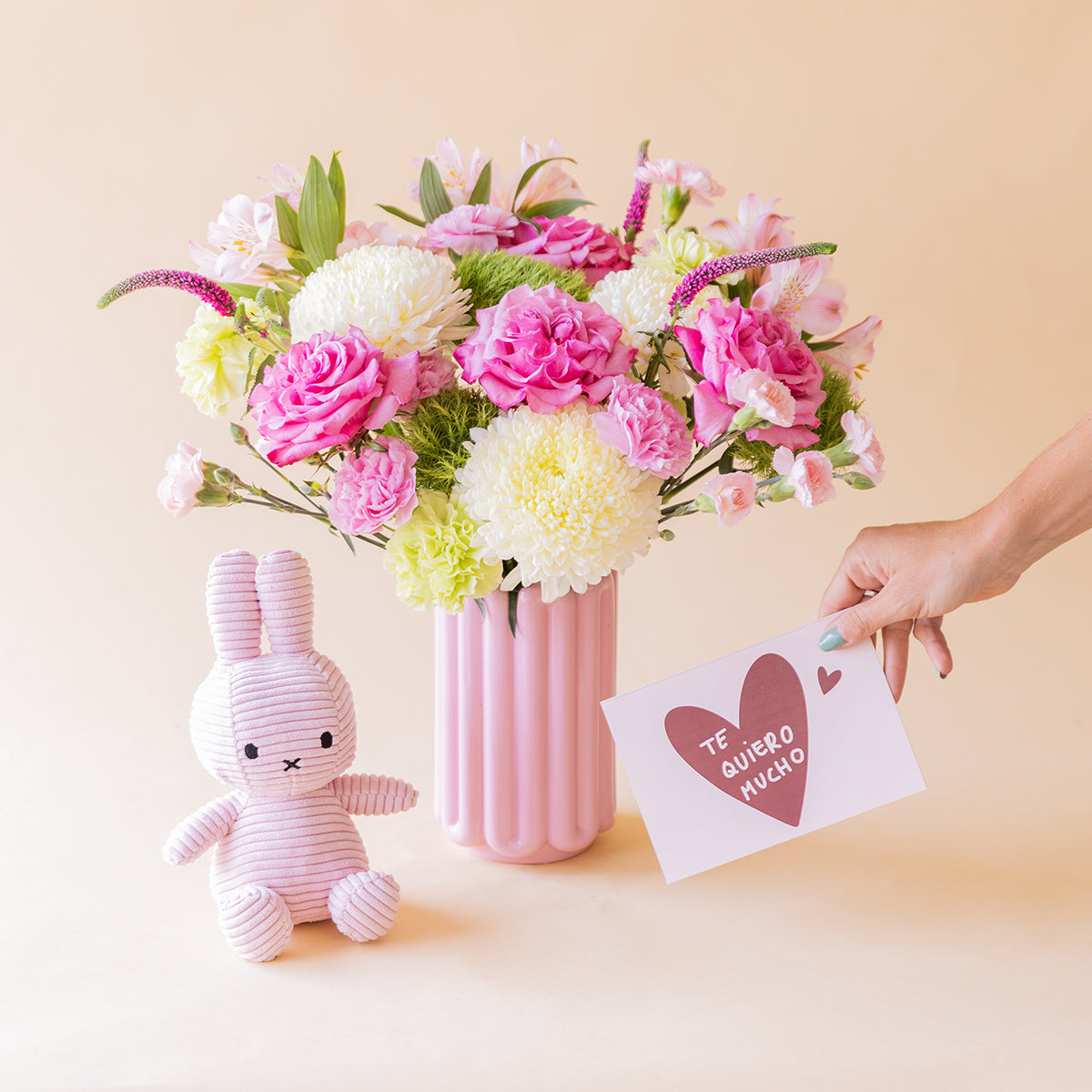 Ramo de rosas fucsias, crisantemos blancos, alstroemeria rosa y veronica lila de Colvin en jarrón acanalado rosa, con peluche Miffy lila y mano sosteniendo tarjeta Te quiero mucho, sobre fondo melocotón