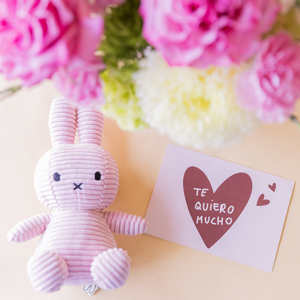 Peluche Miffy de corduroy lila y tarjeta Te quiero mucho con corazón, con ramo de rosas fucsias y crisantemos blancos de Colvin al fondo