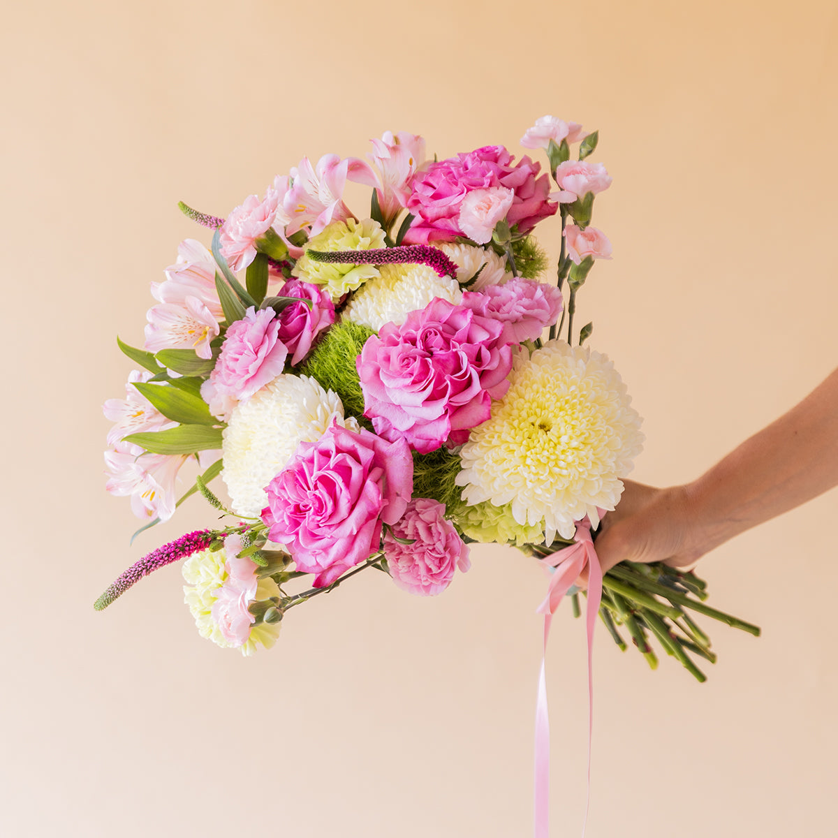 Mano sosteniendo ramo de rosas fucsias, crisantemos blancos, claveles rosas y veronica lila de Colvin, atado con cinta rosa, sobre fondo melocotón