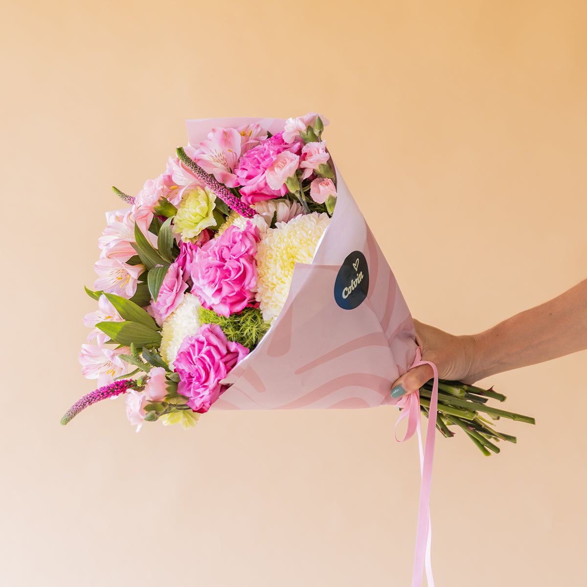 Mano sosteniendo ramo de rosas fucsias y flores rosas de Colvin envuelto en papel rosa con sello Colvin, sobre fondo melocotón