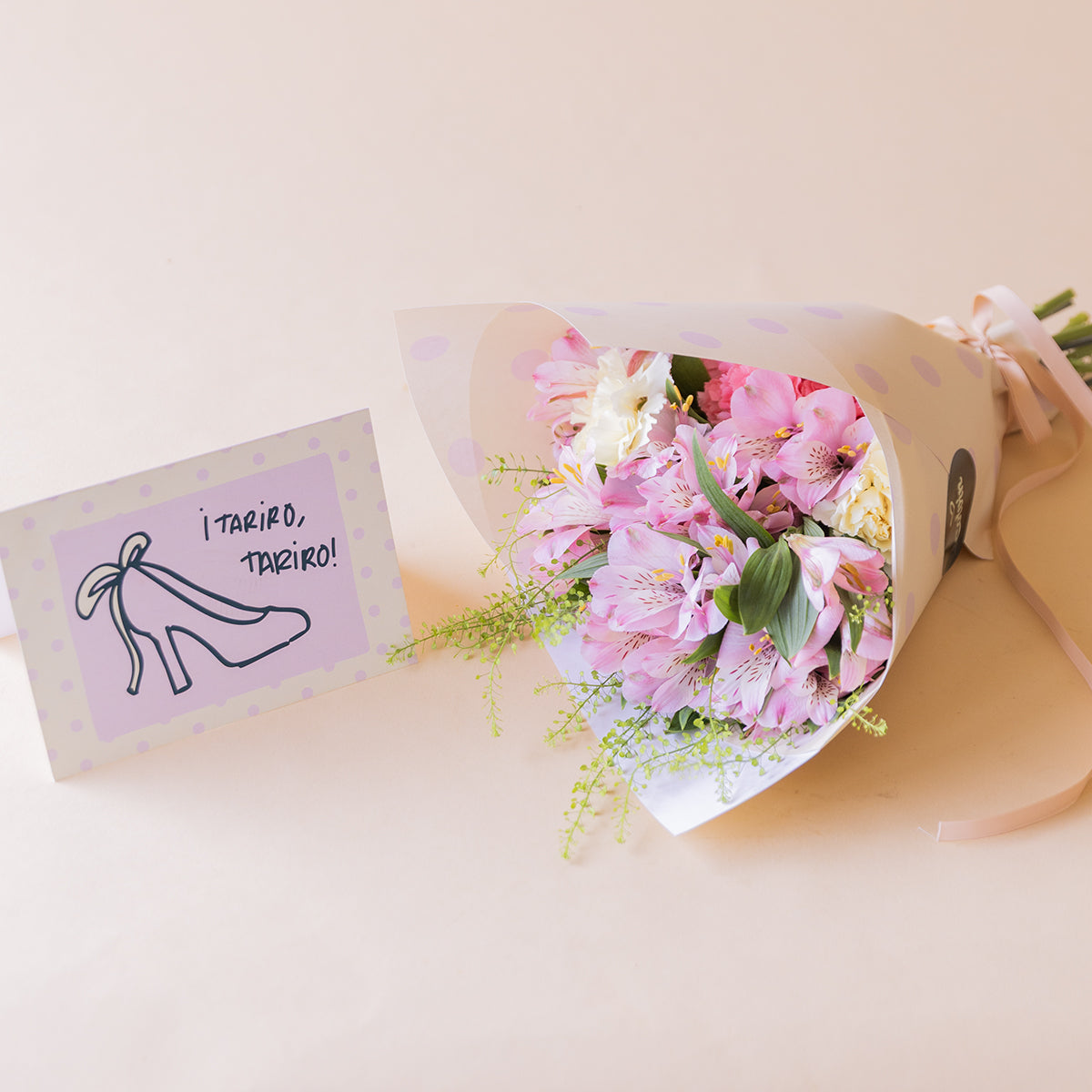 Ramo de alstroemerias rosas, claveles blancos y rosas de Colvin con envoltorio blanco y tarjeta ¡Tariro, Tariro! ilustrada con zapato de tacón, sobre fondo melocotón