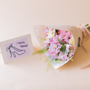 Ramo de alstroemerias rosas, claveles blancos y rosas de Colvin con envoltorio blanco y tarjeta ¡Tariro, Tariro! ilustrada con zapato de tacón, sobre fondo melocotón