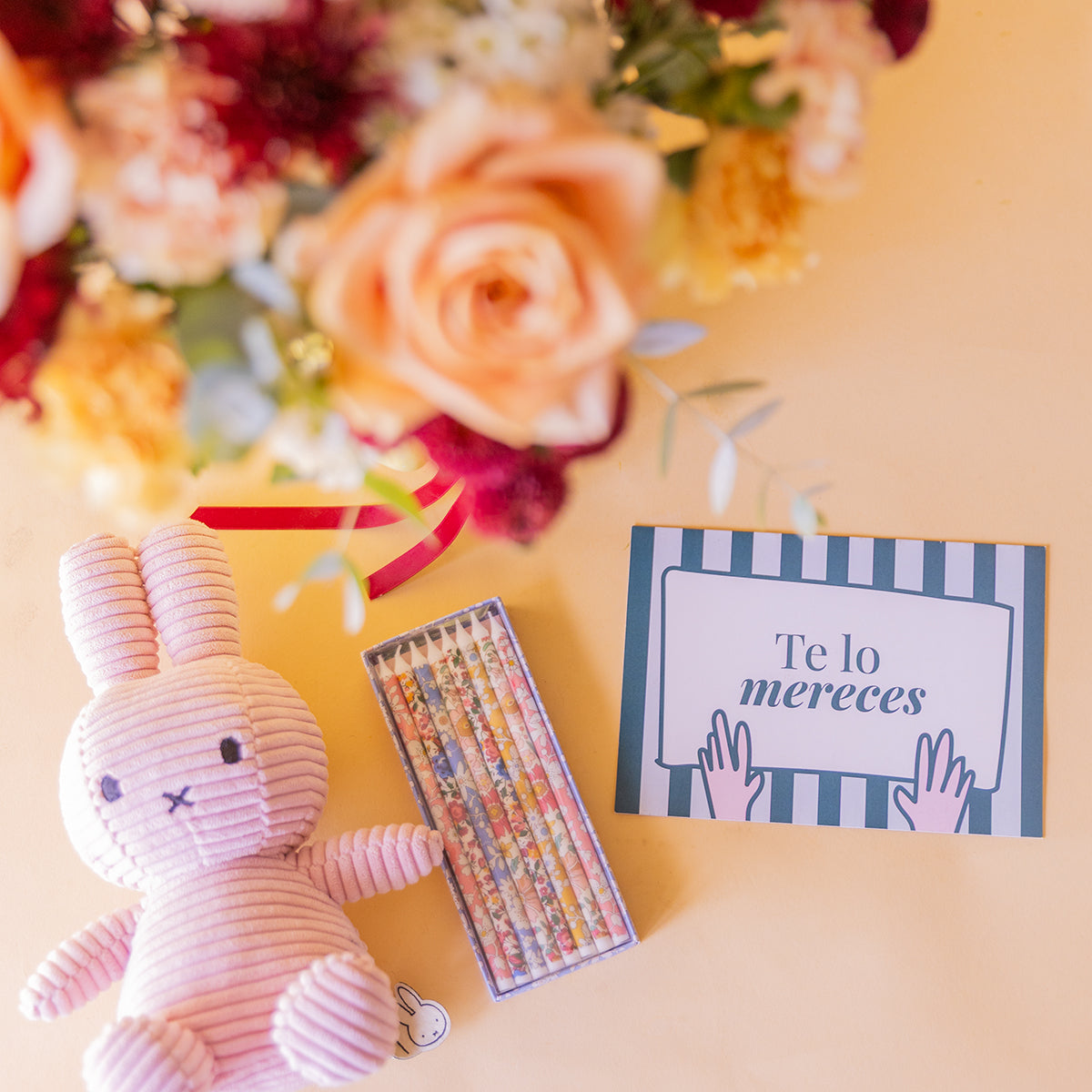 detalle cenital de Miffy rosa, caja de lápices liberty y tarjeta Te Lo Mereces, ramo desenfocado al fondo