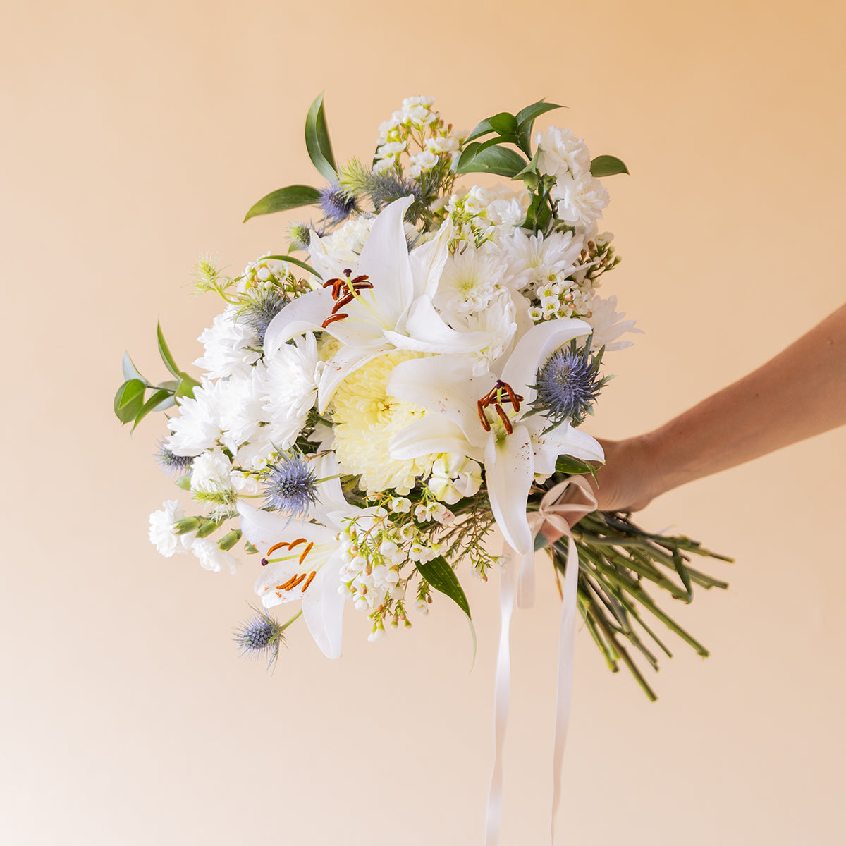Mano sosteniendo ramo de flores blancas de Colvin con lirios, crisantemos, eryngium azul y wax flower, con lazo blanco