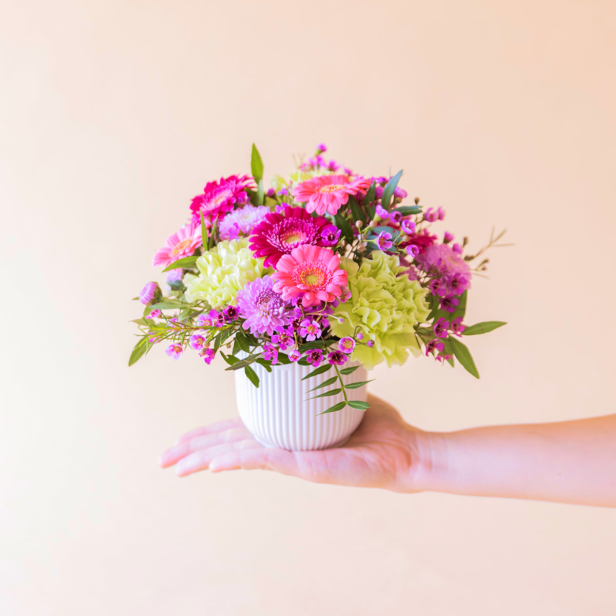 Arreglo floral mini Colvin con gerberas fucsia, claveles verdes y flores silvestres lilas en maceta  estriada.