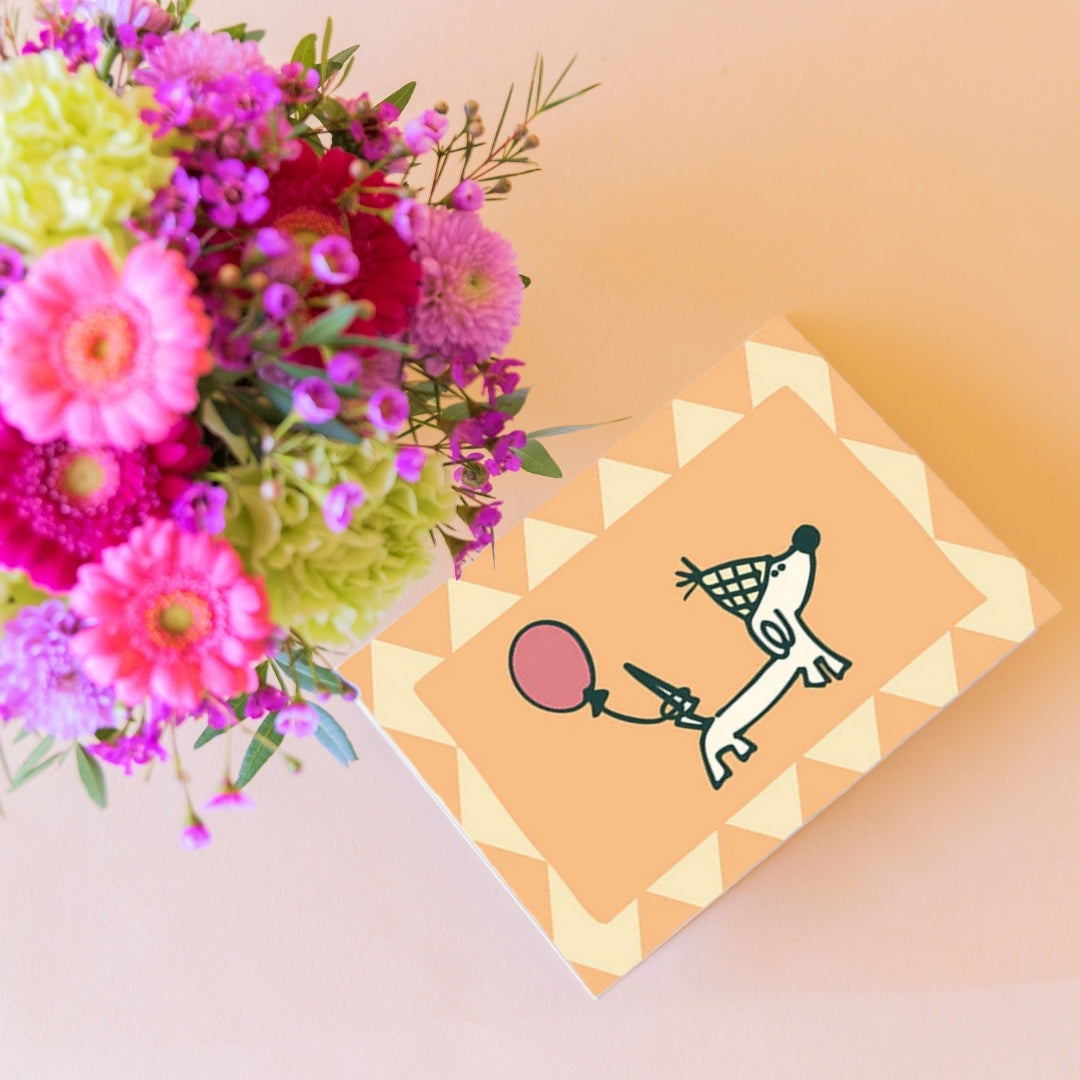 Detalle de tarjeta de cumpleaños Colvin con ilustración de perrito salchicha y globo rosa junto a arreglo floral mini colorido
