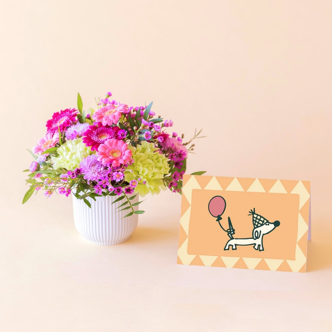 Arreglo floral mini Colvin con flores rosas, lilas y verdes en maceta blanca con tarjeta de cumpleaños con perrito salchicha con globo apoyada al lado