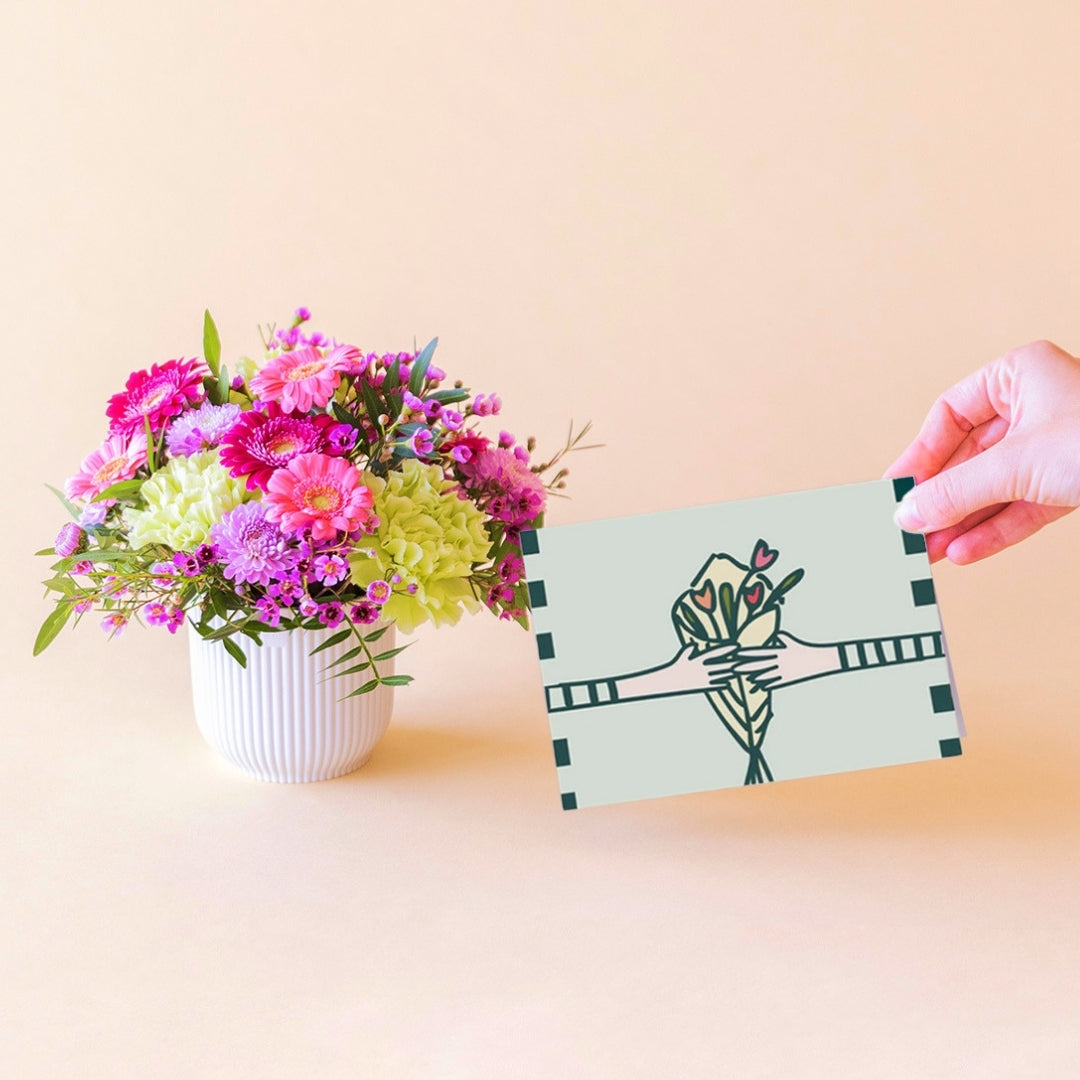 Arreglo floral mini Colvin con gerberas fucsia, crisantemos lilas y claveles verdes en maceta blanca, una mano sosteniendo una tarjeta ilustrada con manos y ramo de flores