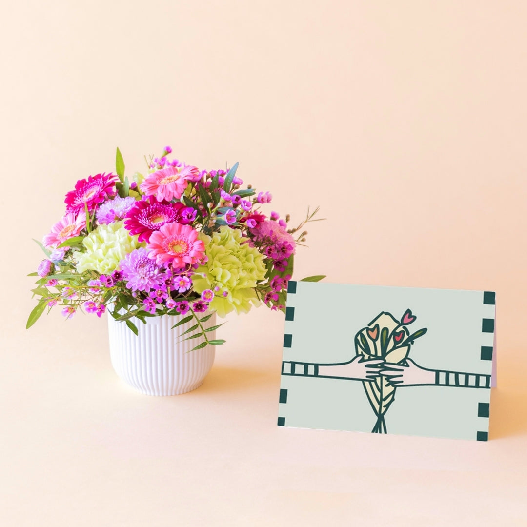 Arreglo floral mini Colvin con gerberas fucsia, crisantemos lilas y claveles verdes en maceta blanca con tarjeta ilustrada apoyada al lado