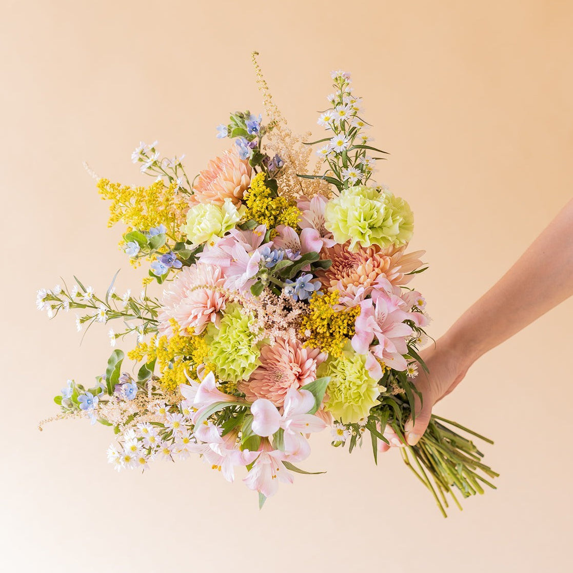 Ramo de flores silvestres Colvin con crisantemos melocotón, alstroemerias y flores amarillas y lilas sobre fondo beige