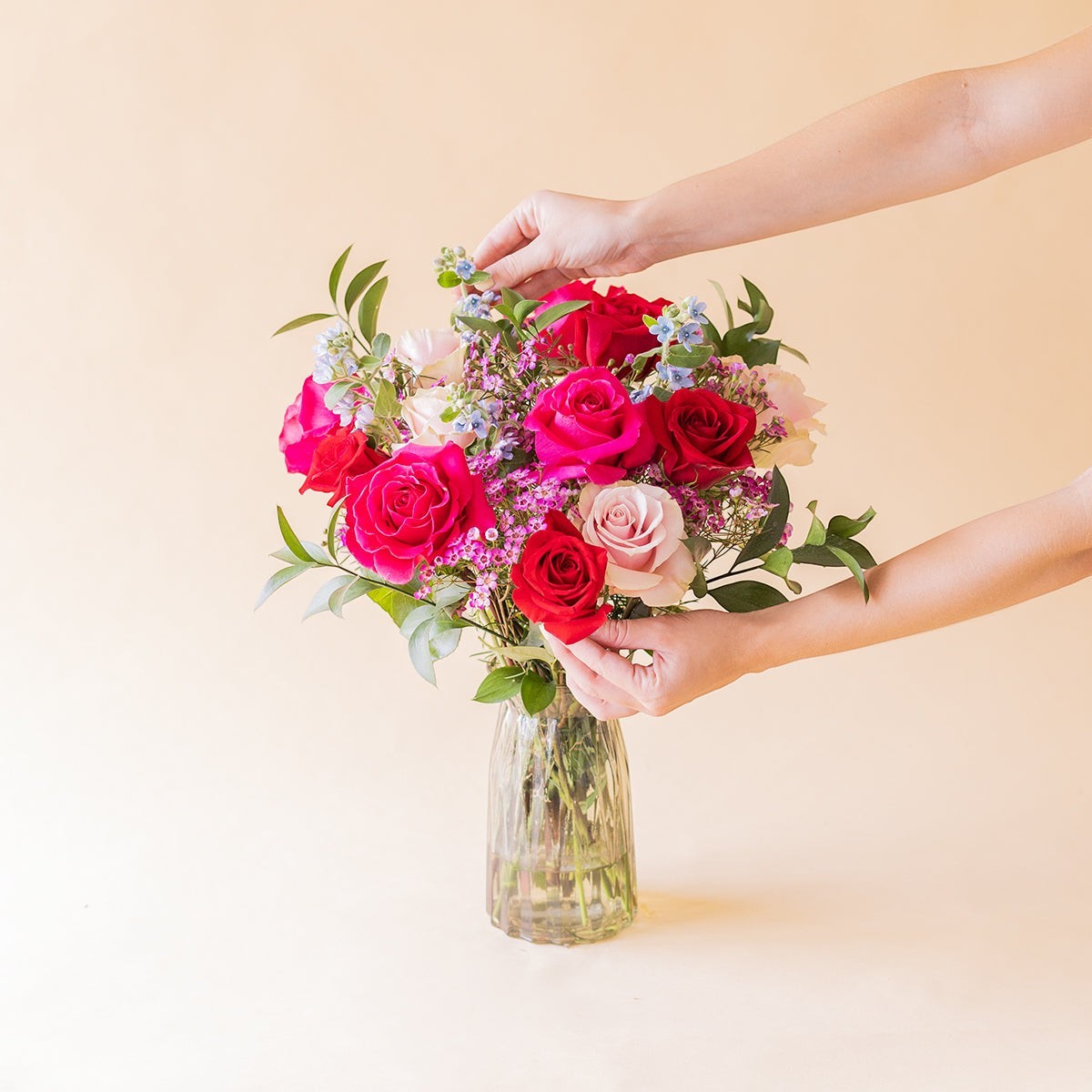 Ramo de rosas fucsias y rojas Colvin en jarrón de cristal, decorado con flores silvestres y follaje verde.