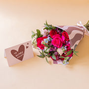 Ramo de rosas fucsias y rojas Colvin con envoltorio de ondas y tarjeta Te quiero mucho sobre fondo beige