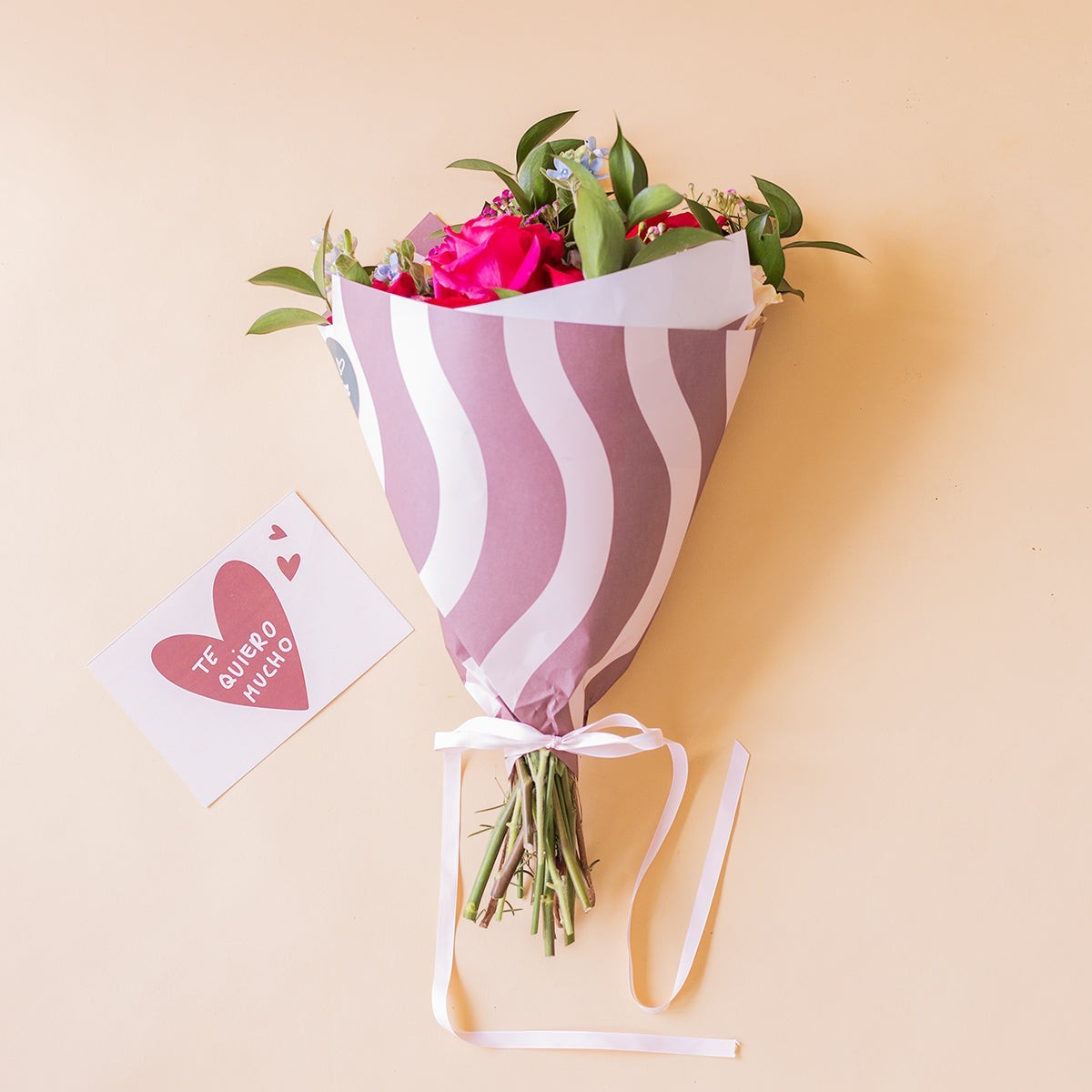 Ramo de rosas fucsia y rojas Colvin con envoltorio de ondas y tarjeta Te quiero mucho sobre fondo beige