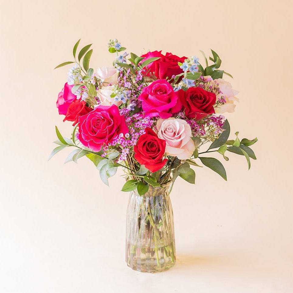 Ramo de rosas multicolores El Encanto de Colvin con flores variadas y follaje verde en un jarrón de cristal