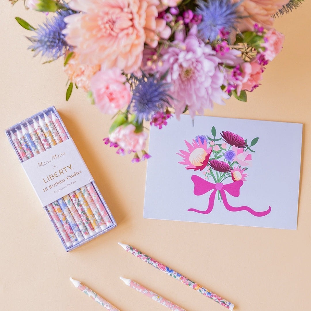 Regalo Colvin con ramo de flores moradas, velas Meri Meri x Liberty estampadas y tarjeta ilustrada sobre fondo beige