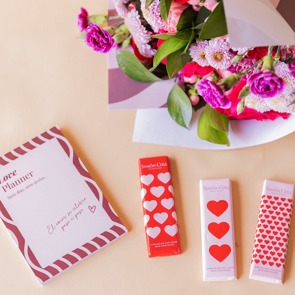 Love Planner de Colvin junto a tres tabletas de chocolate Simón Coll con corazones y ramo de flores fucsia y lila desenfocado