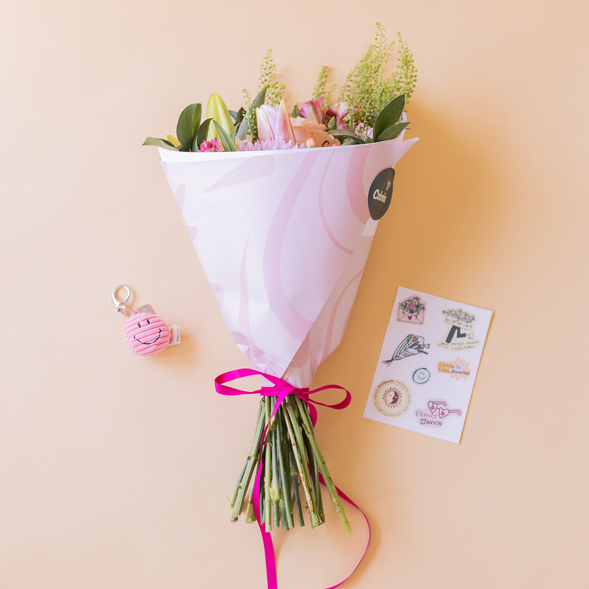 Ramo de flores pastel de Colvin envuelto en papel blanco con lazo fucsia, junto a llavero smiley rosa y hoja de stickers
