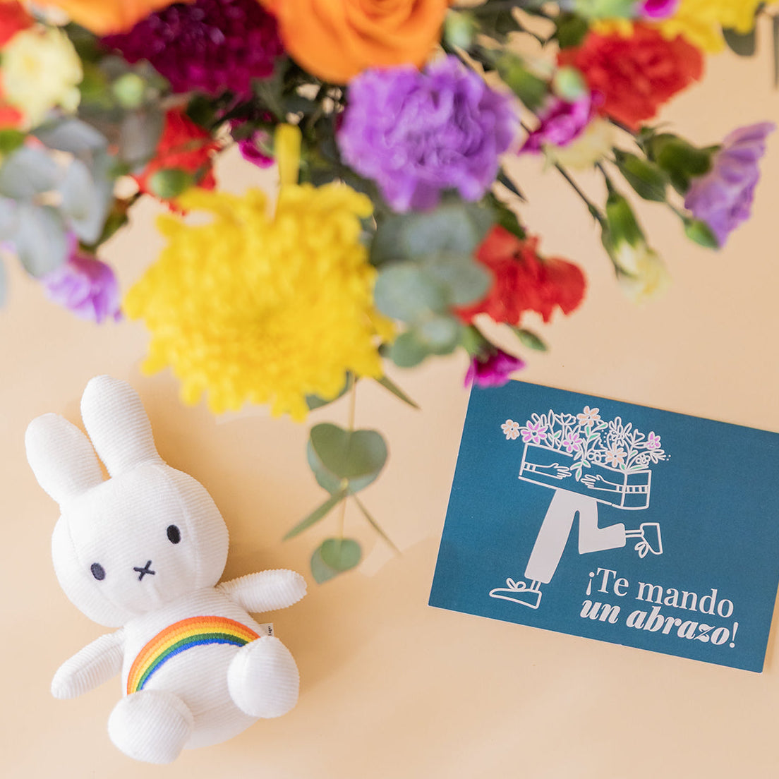 Detalle de peluche Miffy arcoíris y tarjeta azul "¡Te mando un abrazo!" junto a ramo de flores coloridas de Colvin con crisantemos amarillos, rosas naranjas y claveles fucsia