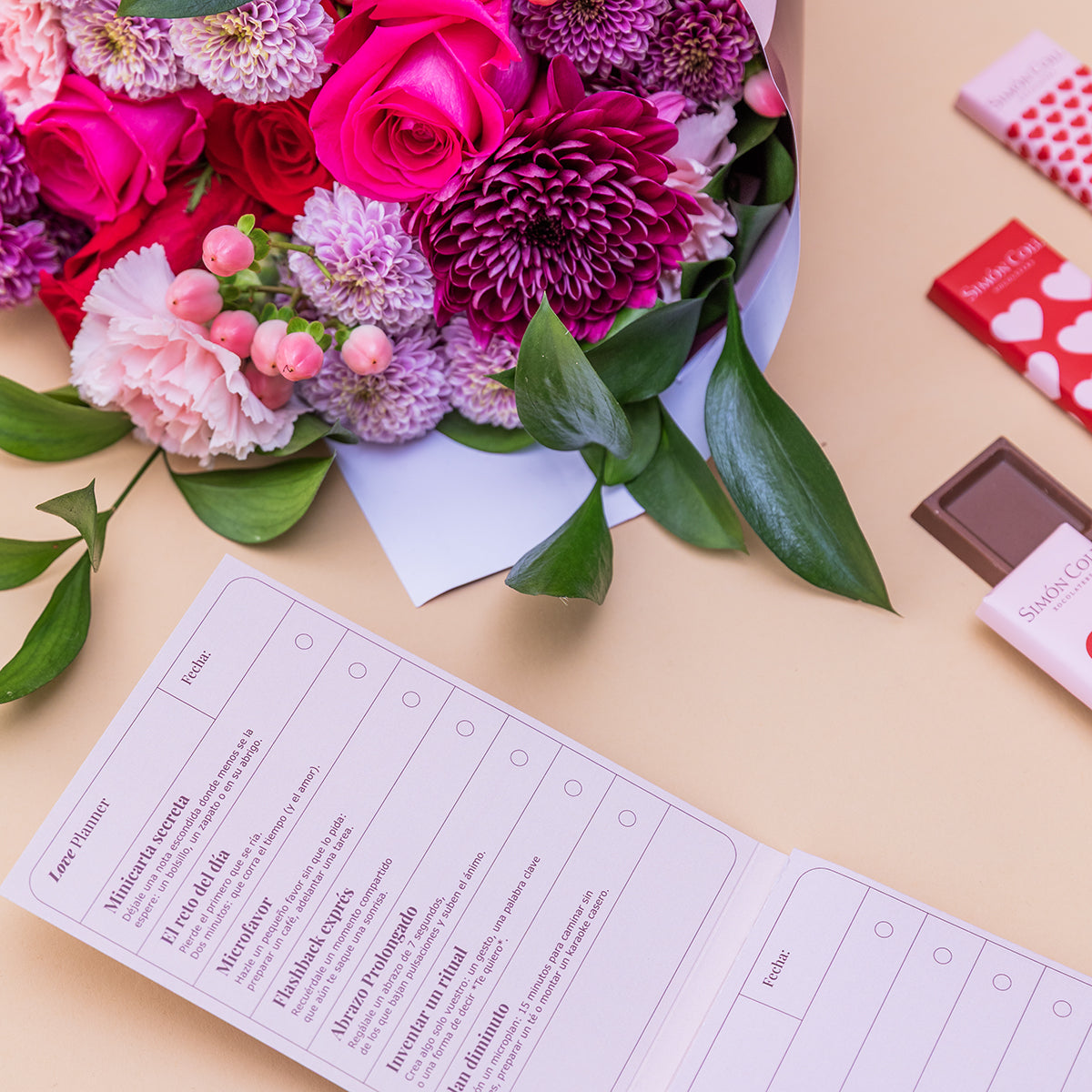 Ramo de flores fucsia y lila de Colvin con Love Planner abierto y chocolates Simón Coll sobre fondo melocotón