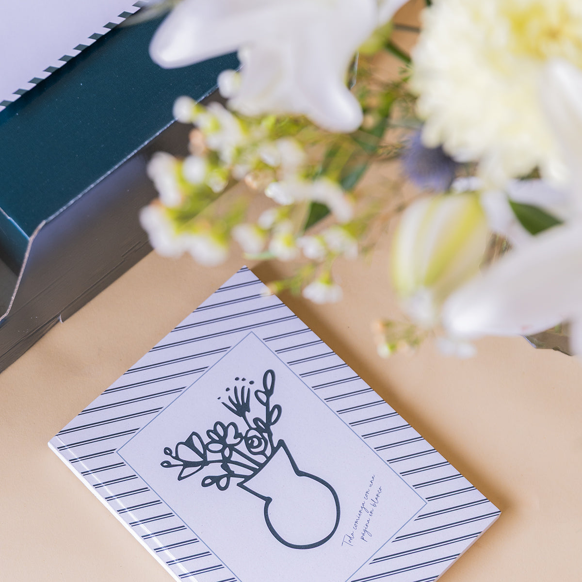 Libreta a rayas de Colvin con ilustración de jarrón de flores, junto a ramo de flores blancas desenfocado