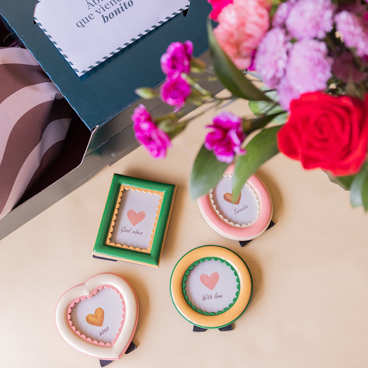 Cuatro marcos de fotos decorativos de Colvin con mensajes junto a caja de envío "Abre, que viene bonito" y ramo de flores rojas y fucsia