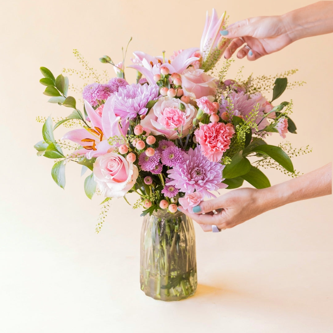 Primer plano del ramo Bonito de Colvin con rosas rosas, claveles rosas, hipericum y crisantemos lila, con dos manos añadiendo flor en el ramo.