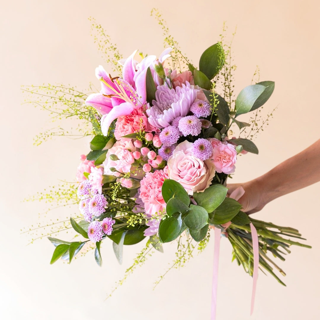 Mano sosteniendo el ramo El Bonito de flores rosas con lirios, crisantemos e hipericum de Colvin