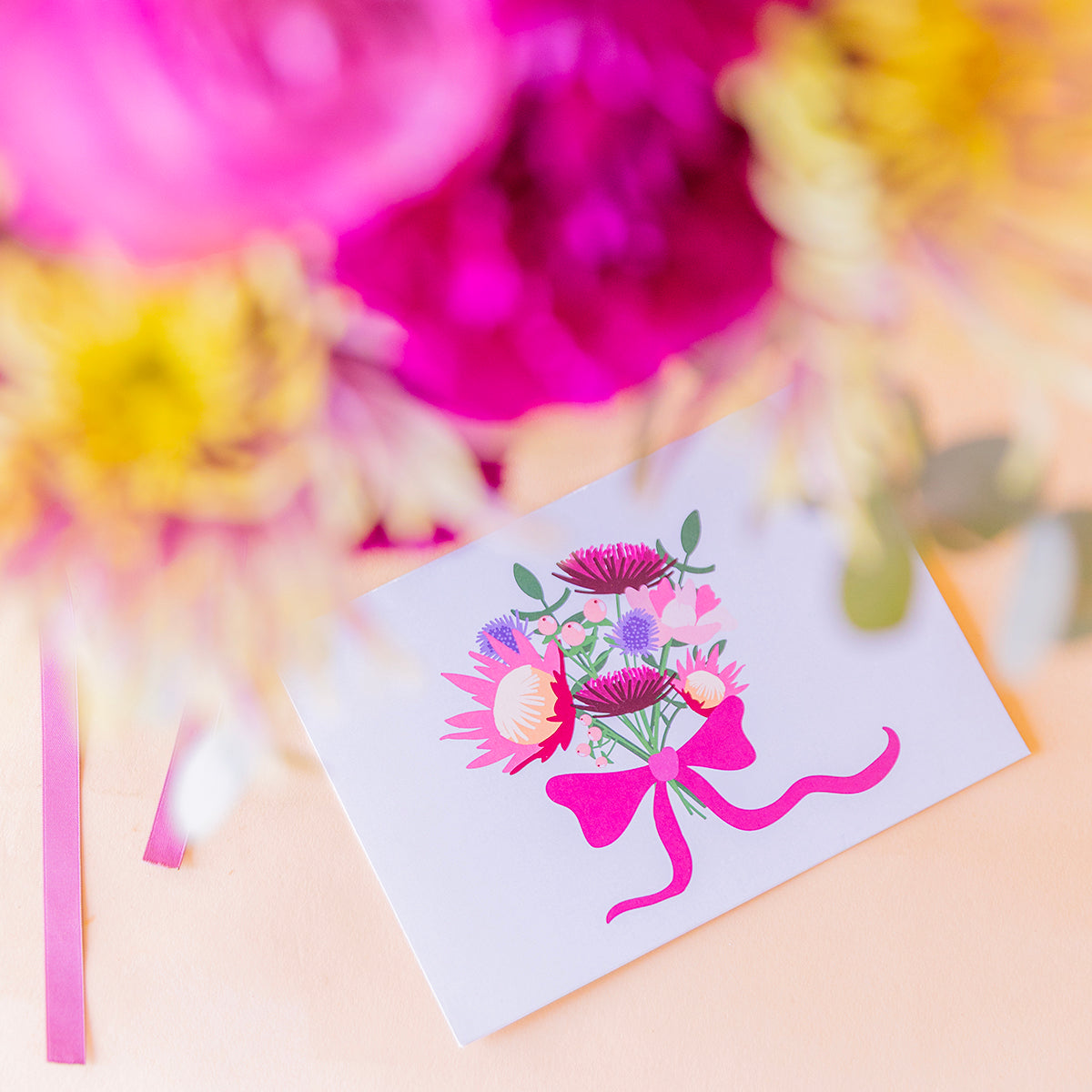 detalle macro de la tarjeta Protea con ilustración de ramo y lazo fucsia, flores desenfocadas al fondo)