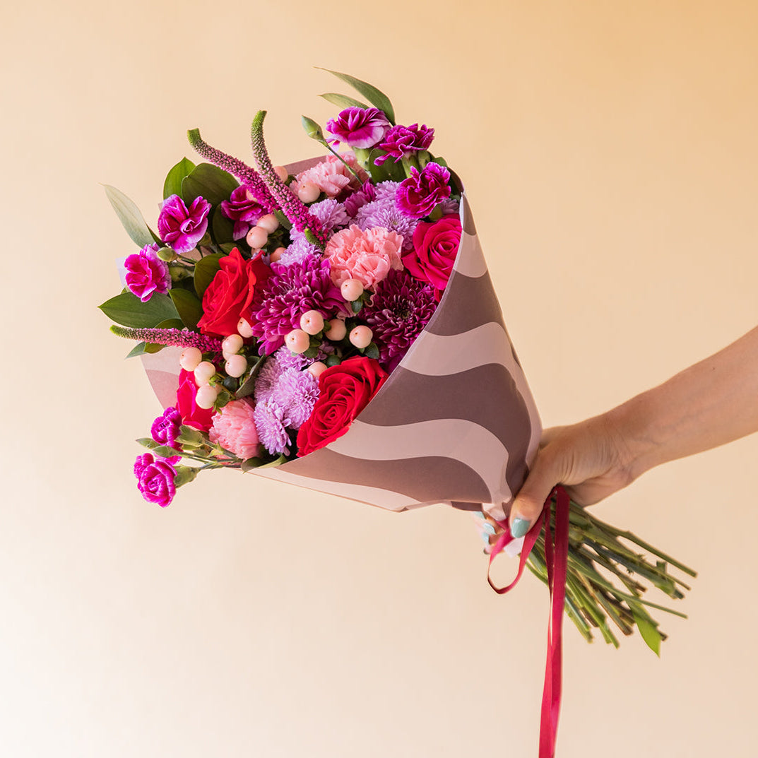 Mano sosteniendo ramo de flores Love is in the Air de Colvin con rosas rojas, crisantemos lilas, claveles fucsia y bayas blancas, envuelto en papel ondulado rosa