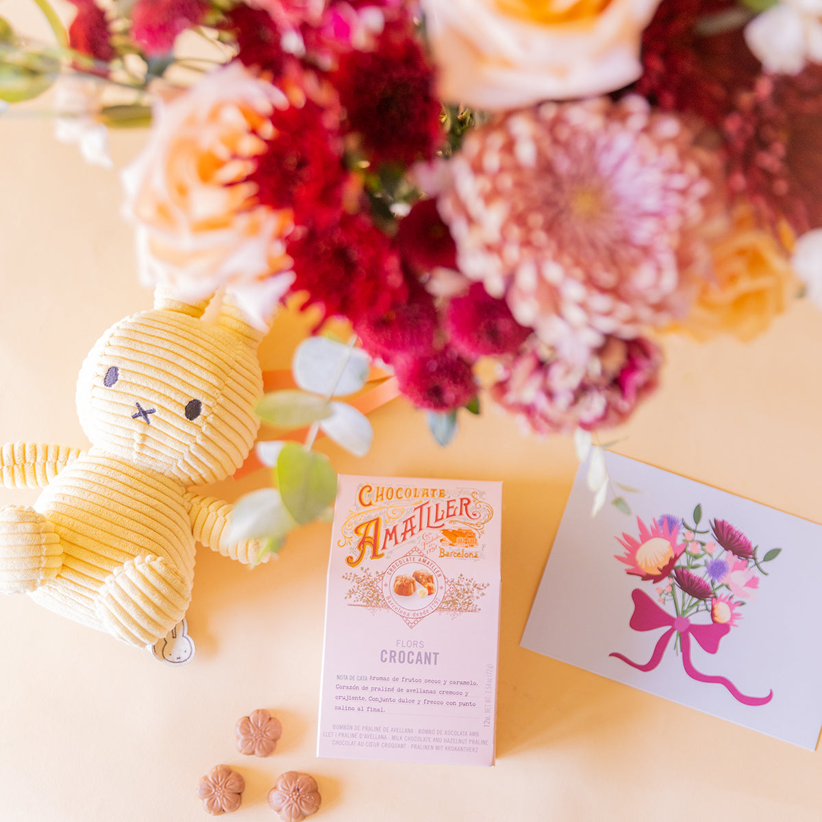 detalle cenital de Miffy amarillo, caja Amatller Crocant y tarjeta Protea, ramo desenfocado al fondo