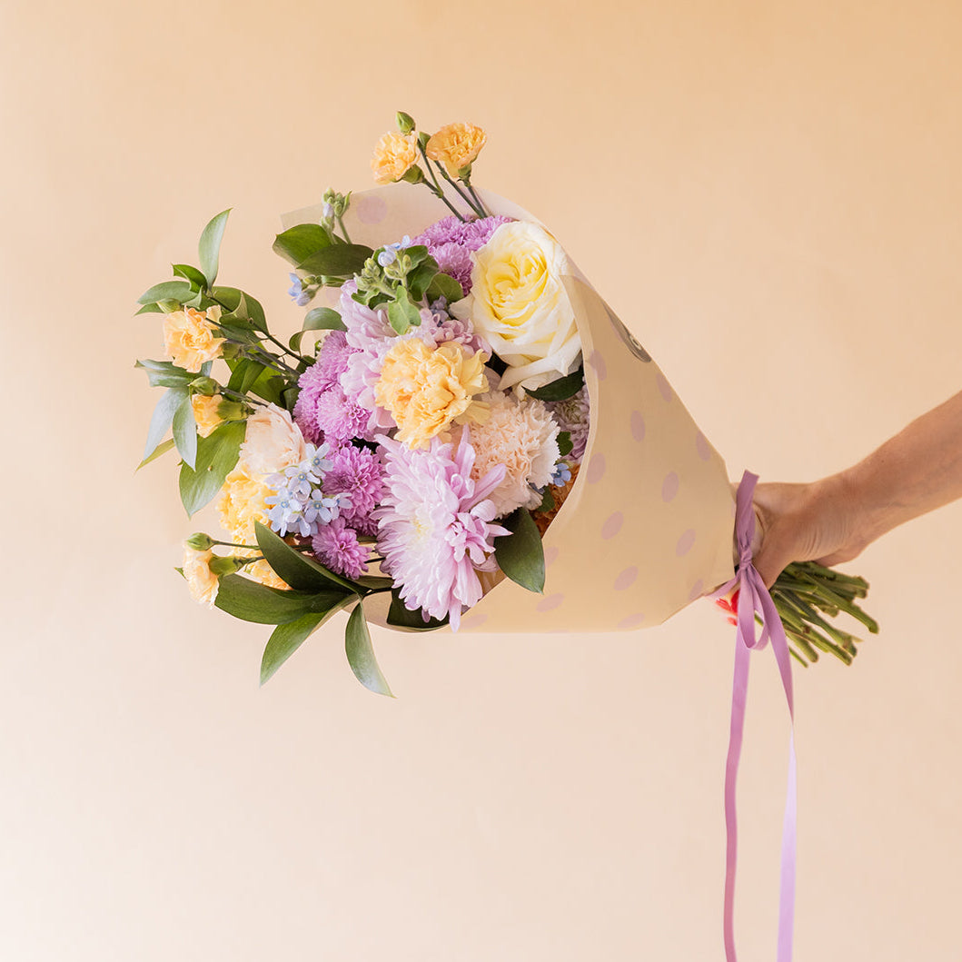 Mano sosteniendo ramo de flores pastel de Colvin con crisantemos lilas, rosas blancas y claveles amarillos envuelto en papel